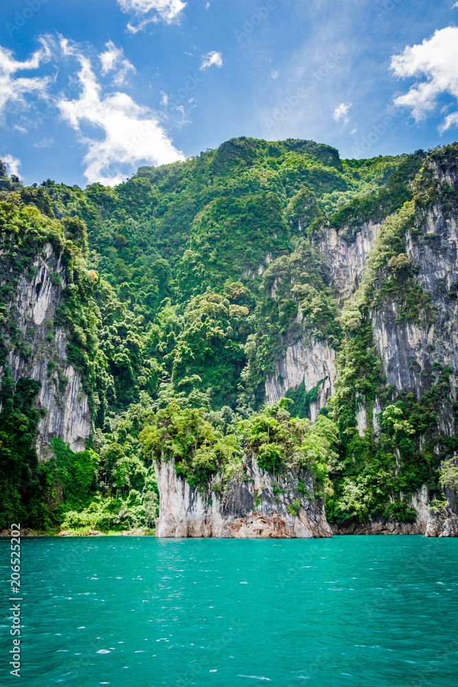 Mountain scenery with with tropical rain forest in the background and blue water lake in the foreground during a sunny day at Ratchaprapha Dam at Khao Sok National Park, Surat Thani Province, Thailand Mountain scenery with with tropical rain forest in the background and blue water lake in the foreground during a sunny day at Ratchaprapha Dam at Khao Sok National Park, Surat Thani Province, Thailand