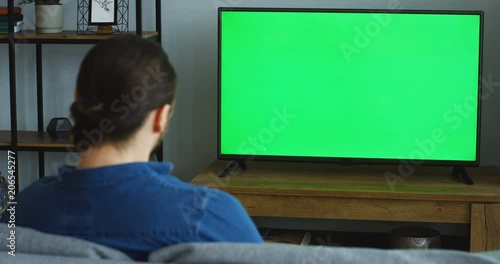 Rear view on the Caucasian man in the jeans blue shirt resting in the living room and turning on TV with green screen with a remote control in his hand. Chroma key. Indoor