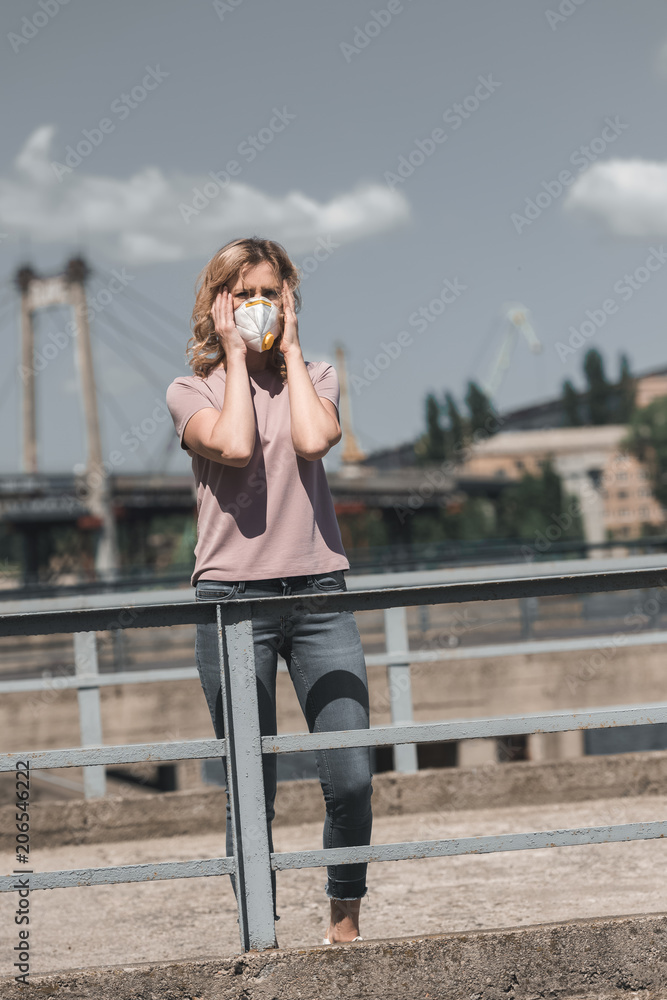 woman in protective mask standing on bridge and touching head, air pollution concept