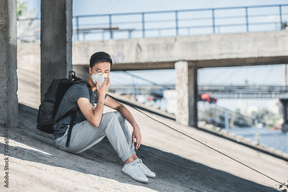 asian teen in protective mask sitting under bridge, air pollution concept