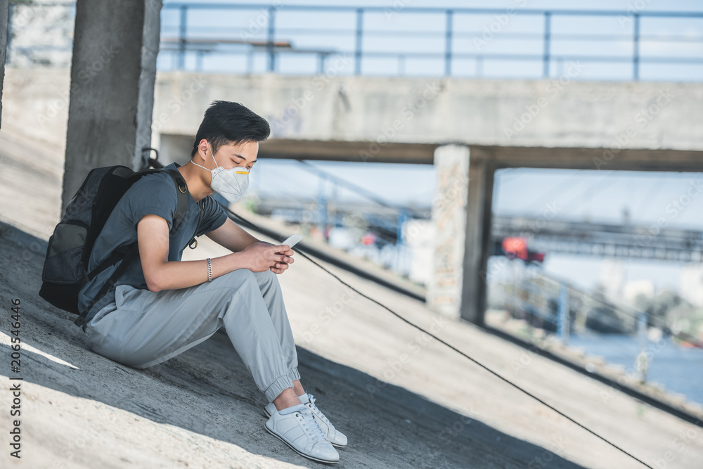 side view of asian teen in protective mask sitting under bridge and using smartphone, air pollution concept side view of asian teen in protective mask sitting under bridge and using smartphone, air pollution concept