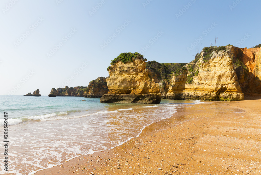 Atlantic sandy beach at sunrise with seashells left by the tide. Waves are rolling among the cliffs under the morning sun in Lagos, Portugal.