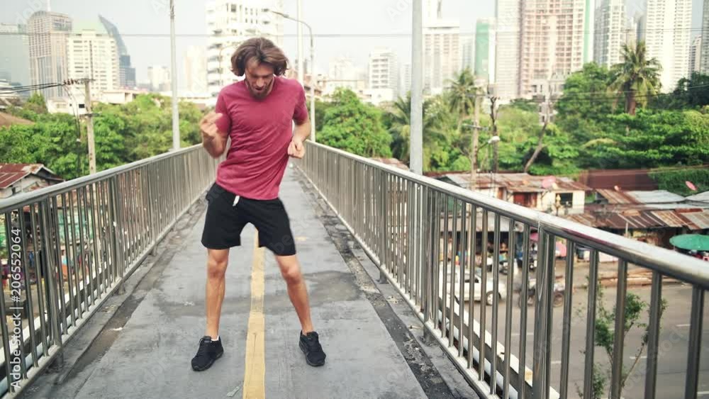 Young LGBT man dancing in urban street. Very happy white male dancing like crazy walking to camera on a overpass with urban city background. Young LGBT man dancing in urban street. Very happy white male dancing like crazy walking to camera on a overpass with urban city background.