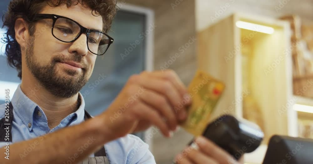 Close up of the handsome man in glasses taking a credit card from female buyer and withdrawing money for buying. Inside Close up of the handsome man in glasses taking a credit card from female buyer and withdrawing money for buying. Inside