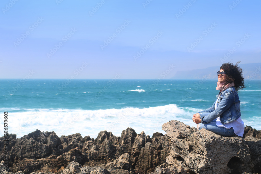Beautiful curly girl sitting on a rock on the ocean background