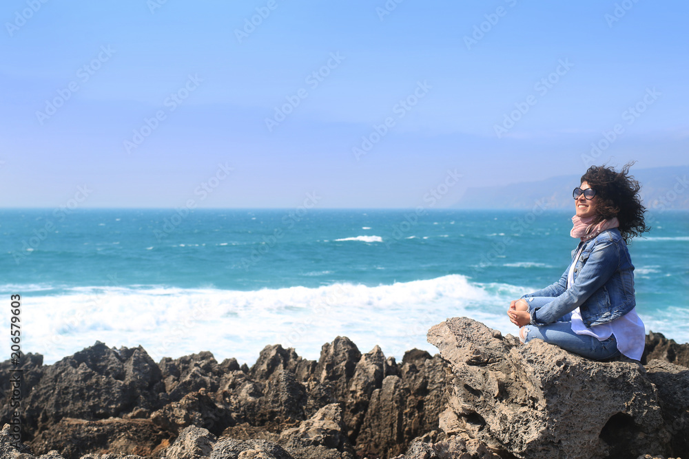 Beautiful curly girl sitting on a rock on the ocean background Beautiful curly girl sitting on a rock on the ocean background