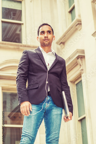 Young Hispanic American Businessman thinking outside in New York, wearing black blazer, blue jeans, white wired earphone, holding laptop computer, standing outside old office building with windows..