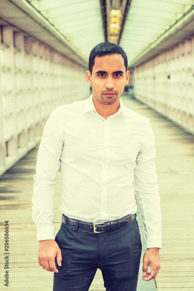 Portrait of Young Handsome Hispanic American Man in New York, wearing white shirt, black pants, standing on walkway with glass wall inside office building, looking at you.. Portrait of Young Handsome Hispanic American Man in New York, wearing white shirt, black pants, standing on walkway with glass wall inside office building, looking at you..