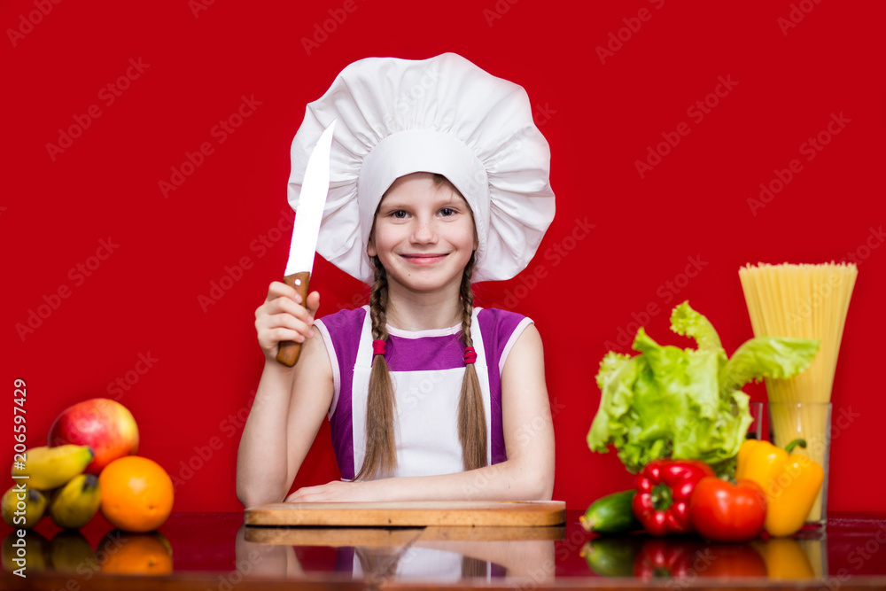Happy little girl in chef uniform cuts fruit in kitchen. Kid chef. Cooking Process Concept Happy little girl in chef uniform cuts fruit in kitchen. Kid chef. Cooking Process Concept