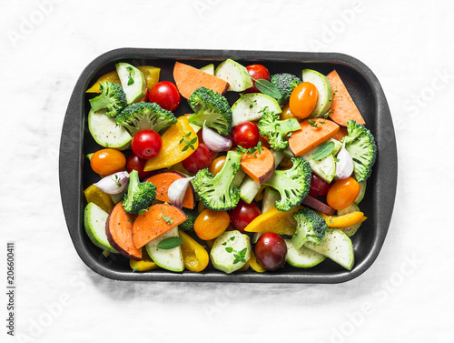 Raw fresh vegetables on a baking sheet. Sweet potato, zucchini, sweet pepper, cherry tomatoes, garlic, broccoli cabbage-ingredients for healthy vegetarian lunch on white background, top view