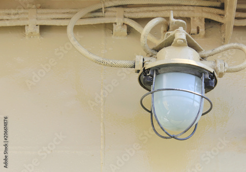 Old Industrial Lamp on Ship Deck Wall with Metal Rustic Frame. Bulkhead Light on Navy Ship against Retro Pale Painted Wall Background. Industrial Simple Light Bulb Lamp Detail Close Up View.