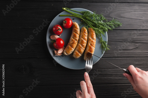 Three grilled sausages on a gray plate on a wooden black background lie also a cherry tomato, greens, garlic, knife, fork. View from above. Place for text and logo. photo