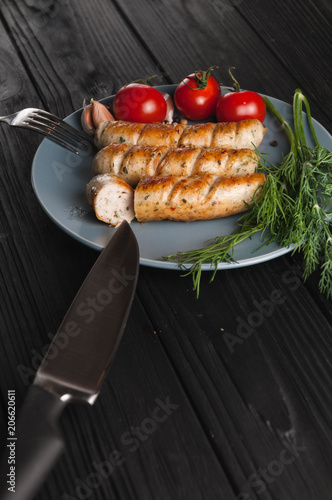 Three grilled sausages on a gray plate on a wooden black background lie also a cherry tomato, greens, garlic, knife, fork. View from above. Place for text and logo. photo