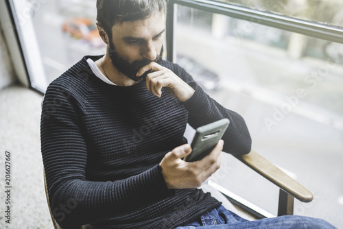 Handsome bearded man using smartphone while resting in modern studio loft. Blurred background photo