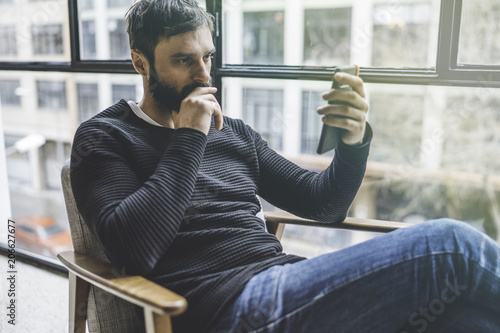 Young cute bearded man sitting home and using smartphone for surfing web internet. Man using gadget at modern apartment. Horizontal. Blurred background photo