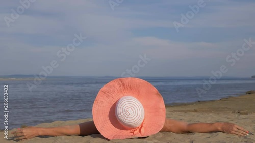 Beautiful woman lies on the beach. Her hands are stretched out to the sides and teasing the sand. She's lying on the beach rug. The girl wears a hat on her head. Nearby lie glasses and slippers. photo