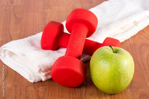 Red fitness dumbbell and green fresh apple with dew on white towel and wooden floor. Diet, sport and health food concept