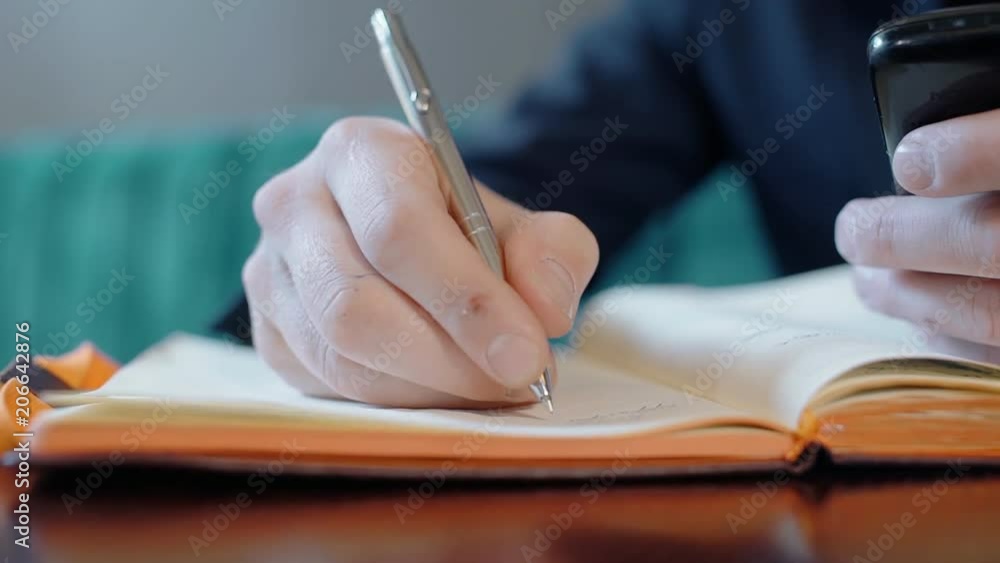Young man is writing in diary, sitting at table in cafe indoors. Successful freelancer works at desk, takes notes in notebook sheets with pen, holding smartphone. Employee in business suit plans Young man is writing in diary, sitting at table in cafe indoors. Successful freelancer works at desk, takes notes in notebook sheets with pen, holding smartphone. Employee in business suit plans