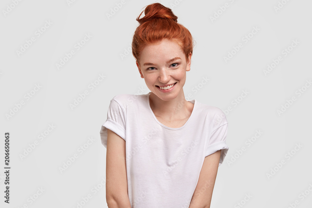 Portrait of happy red haired female being in good mood, wears casual clothing, has shy expression, recieves compliment, stands against white background. Isolated shot. People and skin care concept Portrait of happy red haired female being in good mood, wears casual clothing, has shy expression, recieves compliment, stands against white background. Isolated shot. People and skin care concept