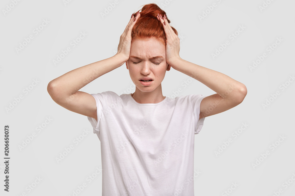 Photo of stressed desperate red haired female has headache, dressed in casual t shirt, feels tired after sleepless night, keeps hands on head, isolated on white concrete wall. Depressed ginger girl Photo of stressed desperate red haired female has headache, dressed in casual t shirt, feels tired after sleepless night, keeps hands on head, isolated on white concrete wall. Depressed ginger girl
