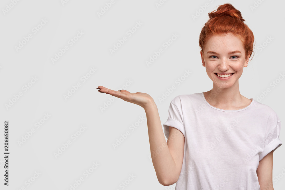 Lovely foxy female demonstrates boutique, keeps palm raised, stands against white background, has positive expression and freckled skin. Pointing at great place, where there is something interesting Lovely foxy female demonstrates boutique, keeps palm raised, stands against white background, has positive expression and freckled skin. Pointing at great place, where there is something interesting