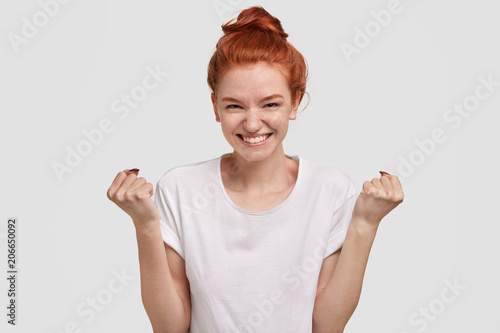 Wallpaper Mural Glad successful ginger female keeps hands in fists, feels her strength, smiles joyfully, celebrates victory, wears white casual t shirt in one tone with background. Monochrome. Victory concept. Torontodigital.ca