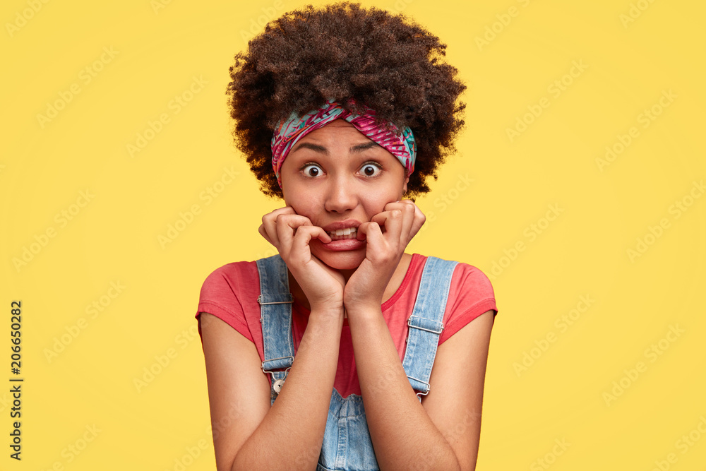 Nervous female with Afro hairstyle, bites finger nails and feels worried, being short of money, has to pay bills, dressed casually, isolated over yellow background. People, emotions concept. Nervous female with Afro hairstyle, bites finger nails and feels worried, being short of money, has to pay bills, dressed casually, isolated over yellow background. People, emotions concept.