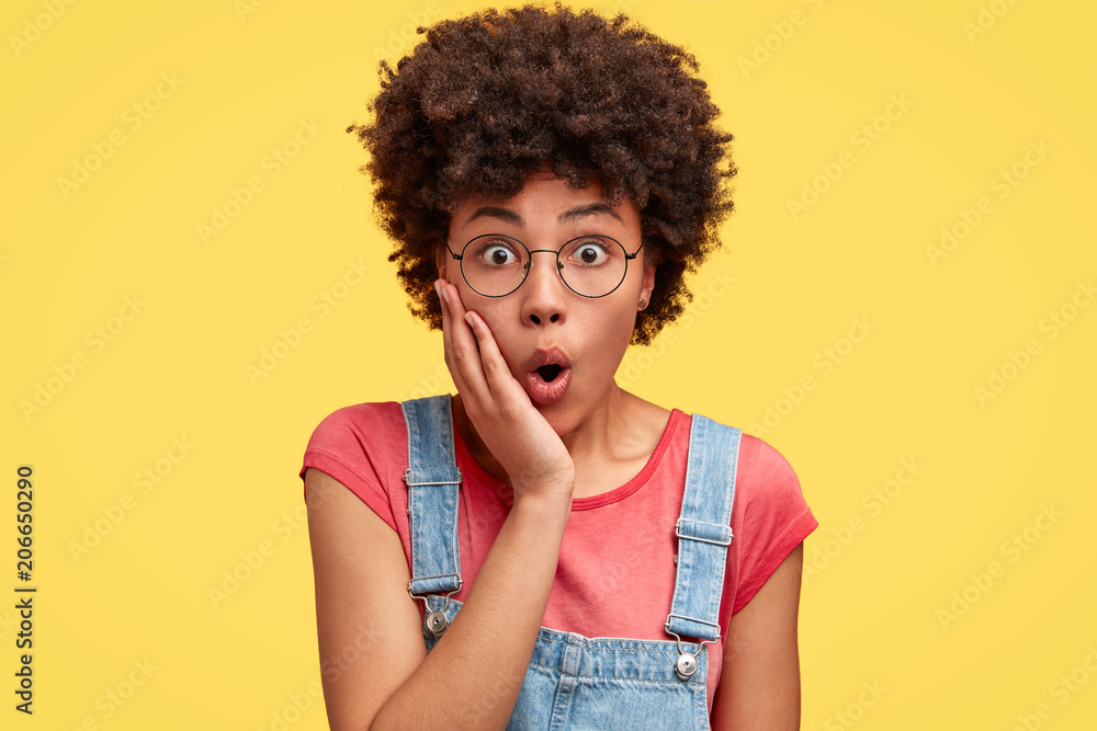 Photo of attractive young female with amazed look, keeps hand on cheek, feels puzzled and surprised as notices something unexpected, has Afro hairstyle, stands indoor against yellow background Photo of attractive young female with amazed look, keeps hand on cheek, feels puzzled and surprised as notices something unexpected, has Afro hairstyle, stands indoor against yellow background