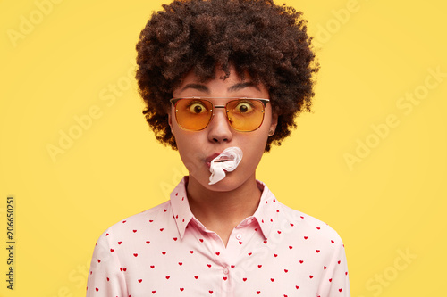 Close up shot of pretty dark skinned African American female blows chewing gum, has stunned expression, forgets to buy something, prepares for party with friends, isolated over yellow background