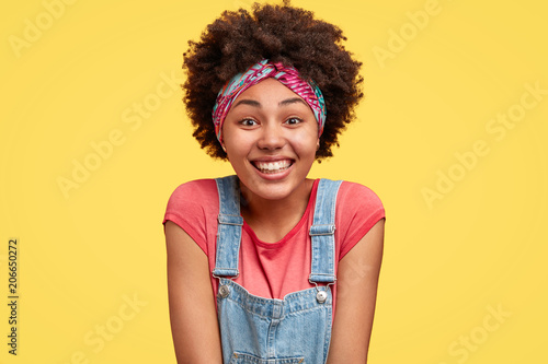 Happy dark skinned young female being in good mood, looks positively at camera, has broad gentle smile, rejoices weekend and free time, dressed in jean overalls, poses against yellow background.