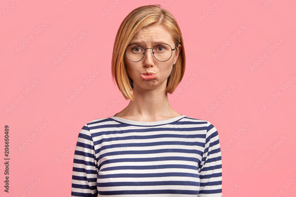 Emotive abused Caucasian female with sorrowful expression, curves lower lips with discontent look, recieves bad words concerning her work, feels frustrated, wears spectacles and striped jacket Emotive abused Caucasian female with sorrowful expression, curves lower lips with discontent look, recieves bad words concerning her work, feels frustrated, wears spectacles and striped jacket