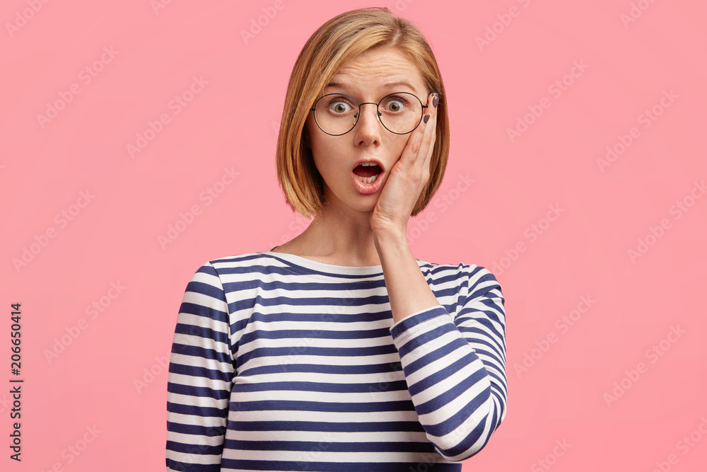 Beautiful amazed Caucasian young female keeps hand on cheek, looks puzzled and shocked, misses train as comes too late on railway station, stares at new schedule, wears striped casual sweater Beautiful amazed Caucasian young female keeps hand on cheek, looks puzzled and shocked, misses train as comes too late on railway station, stares at new schedule, wears striped casual sweater