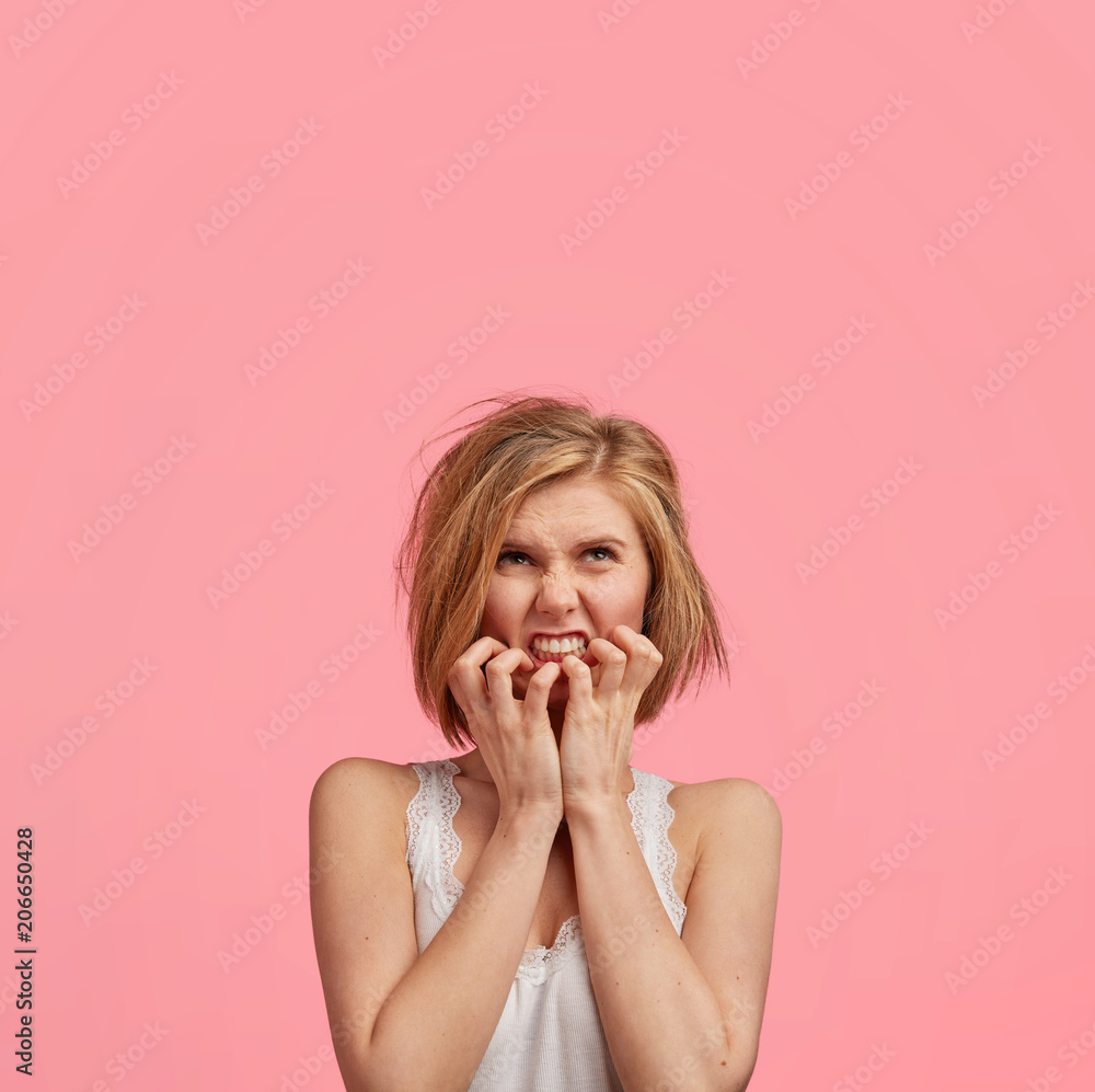 Portrait of nervous female feels puzzled and frustrated, bites finger nails, has mess on head, wears nightwear, poses against pink background with blank space upwards for your promotional text Portrait of nervous female feels puzzled and frustrated, bites finger nails, has mess on head, wears nightwear, poses against pink background with blank space upwards for your promotional text