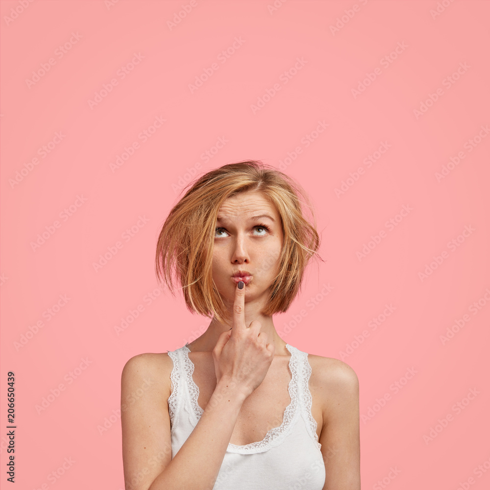 People and thoughts concept. Attractive Caucasian woman looks pensively upwards, keeps fore finger near round lips, wears sleepwear, plans day, has something in mind, isolated on pink studio wall People and thoughts concept. Attractive Caucasian woman looks pensively upwards, keeps fore finger near round lips, wears sleepwear, plans day, has something in mind, isolated on pink studio wall