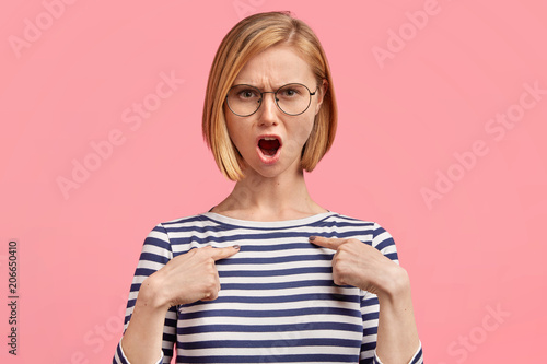 Grumpy annoyed angry female model wears casual sailor jacket and spectacles, indicates at blank space of clothes, dissatified with material, isolated on pink background. Negative human emotions