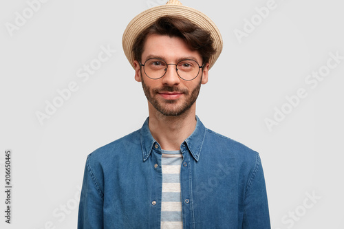 Confident attractive man with stubble, wears denim shirt and hat, being confident and self assured, stands against white background. Stylish traveller ready to explore new places and country