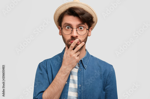 Horizontal portrait of astonished unshaven male keeps hand on mouth, looks in bewildrement, being surprised to hear news from interlocutor, wears starw hat, poses indoor against white concrete wall