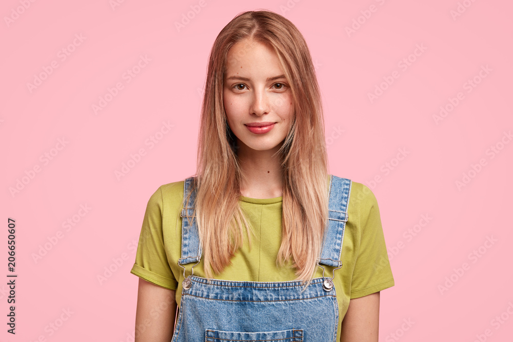 Φωτογραφία Candid shot of good looking Caucasian female with long straight hair, wears jean dungaree, looks confidently at camera, enjoys spare time, going to have walk with her dog outside