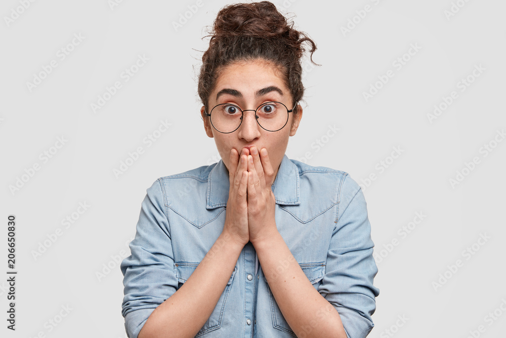 Pretty female waits with temptation for important results, wears denim jacket and spectacles, covers mouth, being excited, loses speech, stands on white background. Teenager watches breathtaking scene Pretty female waits with temptation for important results, wears denim jacket and spectacles, covers mouth, being excited, loses speech, stands on white background. Teenager watches breathtaking scene