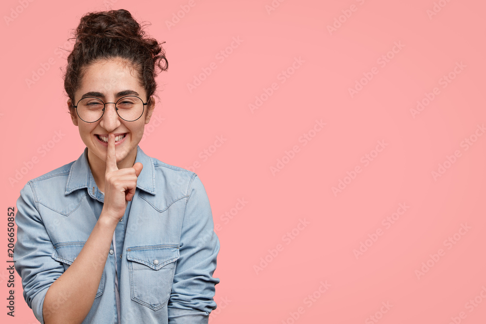Indoor shot of good looking smiling female with dark curly hair, makes hush gesture, giggles at camera, tells secret about her wedding to best friend, stands against pink wall with copy space
