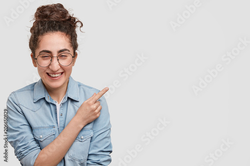 Positive brunette female wonk student has comic expression, giggles at camera with joyful expression, indicates at right upper corner, presents her project or some funny photos on whiteboard photo