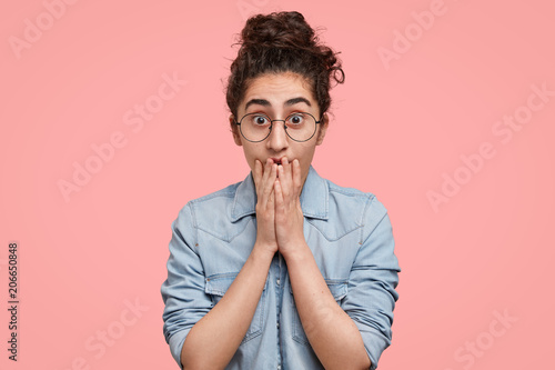 Lovely Cucasian female model sees something unbelievable  covers mouth with hands as expresses great surprisement and confusion  wears casual clothes  poses against pink background. It is insane