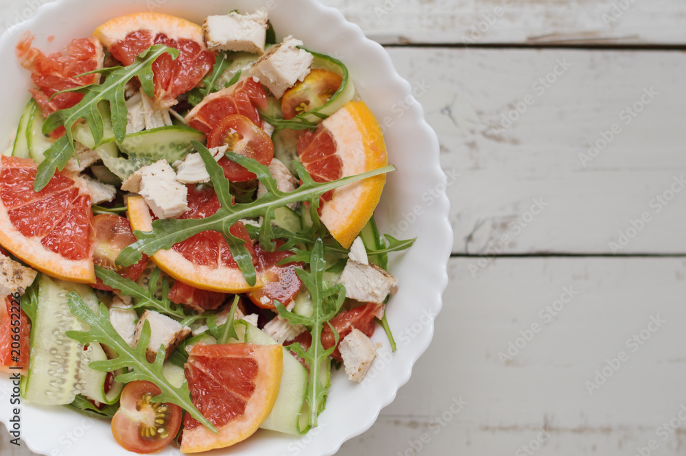 Portion of healthy salad with arugula, grapefruit, cucumber slices, chicken breast, cherry tomatoes in white bowl on gray wooden table, top view
