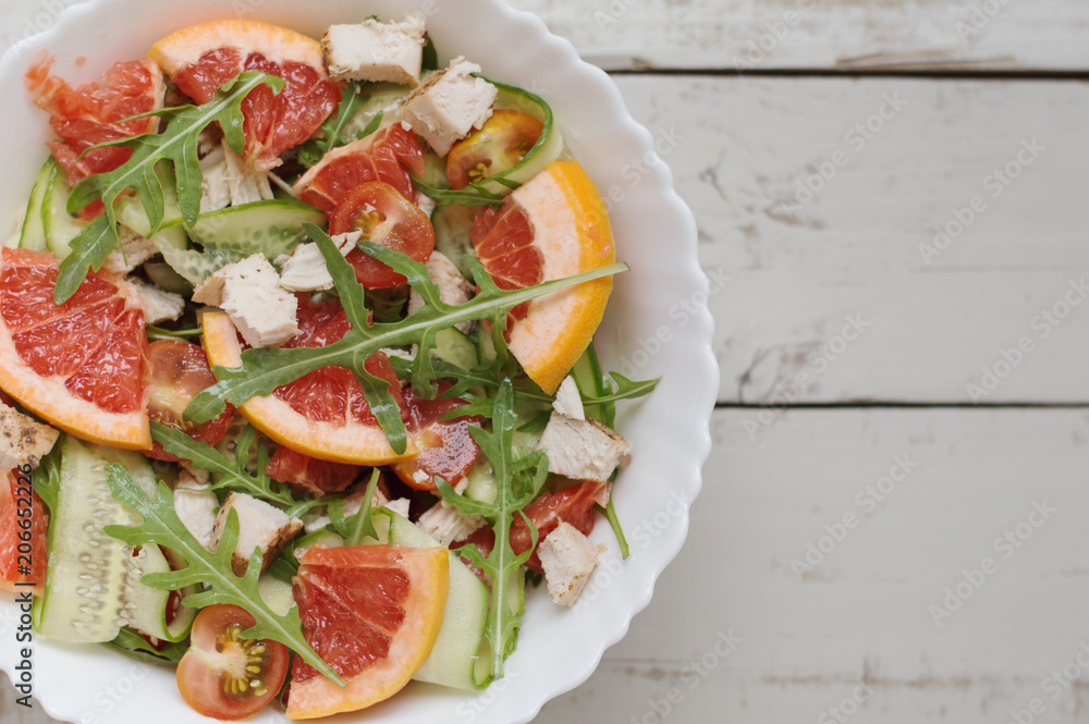 Portion of healthy salad with arugula, grapefruit, cucumber slices, chicken breast, cherry tomatoes in white bowl on gray wooden table, top view Portion of healthy salad with arugula, grapefruit, cucumber slices, chicken breast, cherry tomatoes in white bowl on gray wooden table, top view