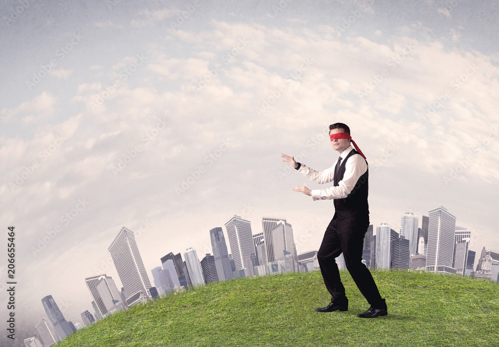 Young blindfolded businessman steps on a a patch of grass with a grey buildings in the background Young blindfolded businessman steps on a a patch of grass with a grey buildings in the background