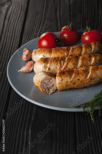 Three grilled sausages on a gray plate on a wooden black background with cherry tomatoes, herbs, garlic.