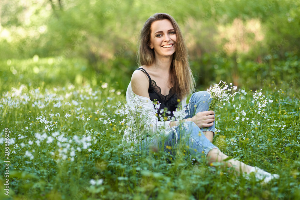 portrait of a happy laughing young woman sitting in the grass on a green meadow among the flowers with a bouquet of small white flowers. portrait of a happy laughing young woman sitting in the grass on a green meadow among the flowers with a bouquet of small white flowers.