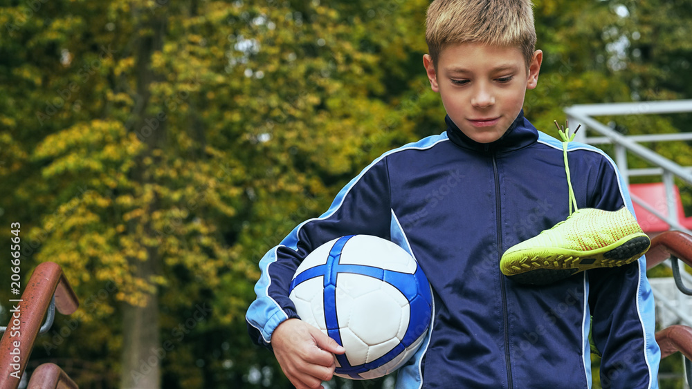 Obraz premium Smiling teenage boy with a soccer ball in his hand and soccer boots on the shoulder against the background of the stadium. Sports training in the field.