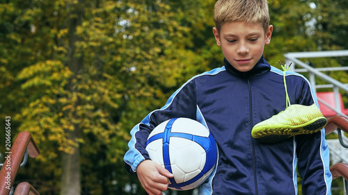 Smiling teenage boy with a soccer ball in his hand and soccer boots on the shoulder against the background of the stadium. Sports training in the field.
