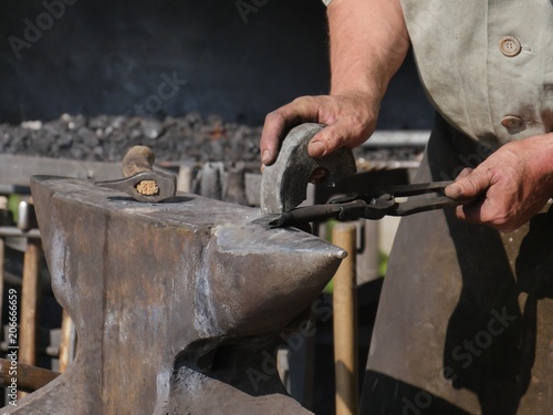 steel anvil, on top of which lies a hot workpiece which is held by a pair of pliers, semi-circular smoothing stone in the hand of the blacksmith, fuzzy background